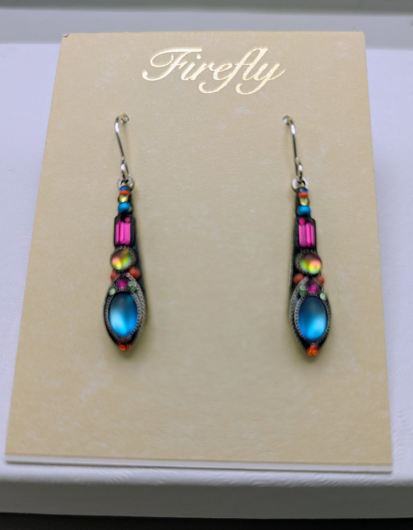A pair of Firefly brand drop earrings with iridescent blue, pink, and orange beads on silver hooks, displayed on a beige card inside a white box.
