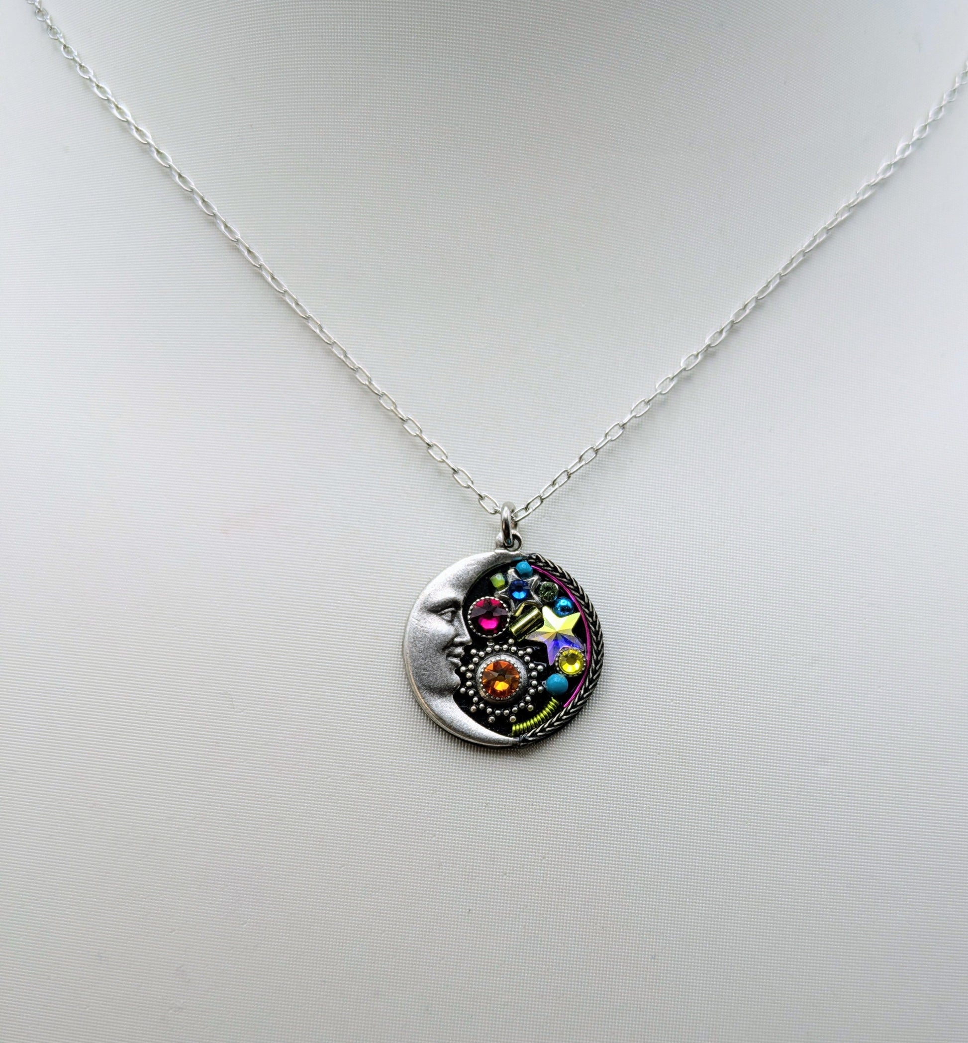 A silver pendant necklace displayed on a white mannequin bust. The pendant is a circular silver disc featuring a colorful celestial design with a crescent moon, stars, and a central sun-like motif with pink, blue, yellow, and orange details.