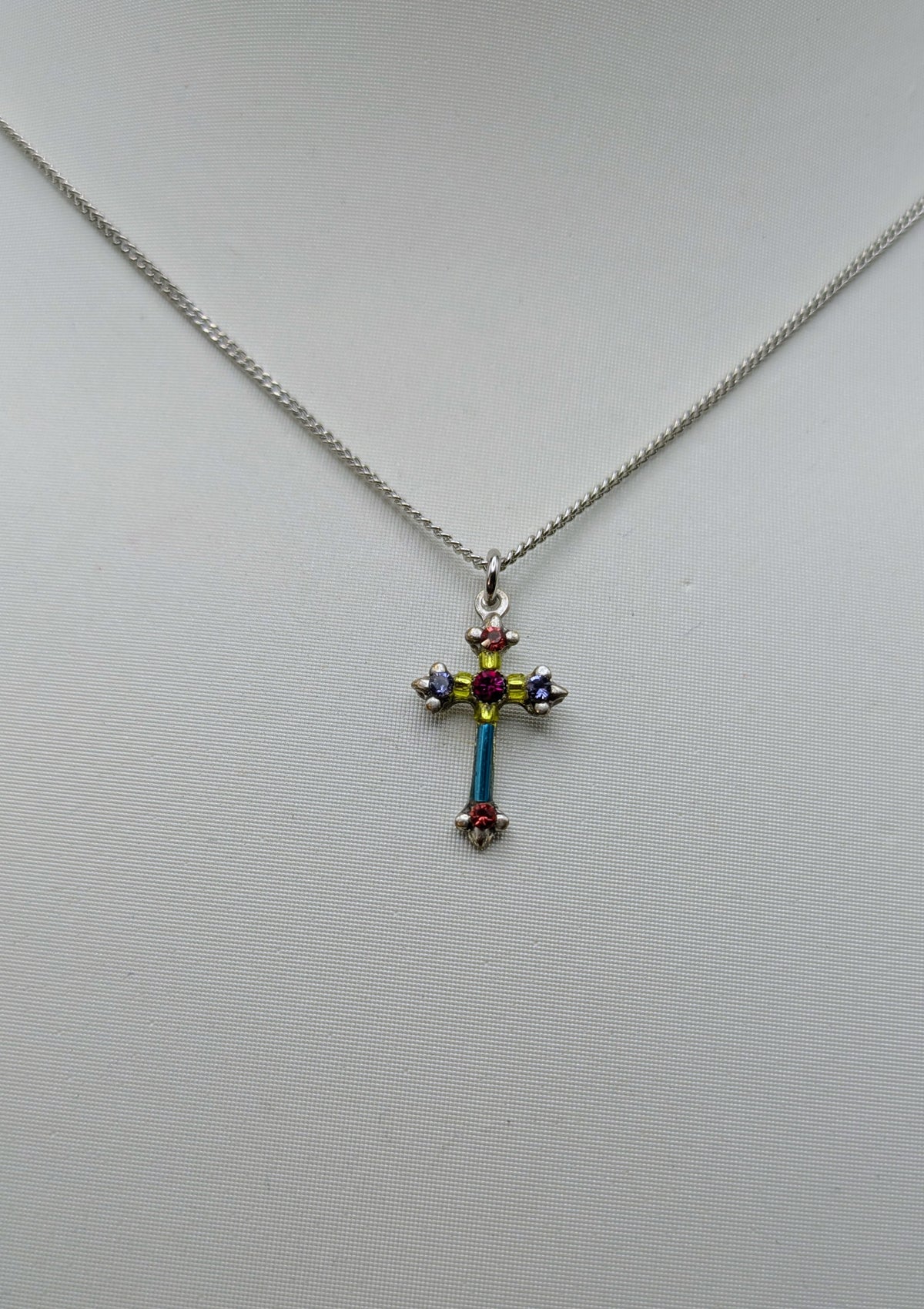 A silver cross pendant necklace with colorful enamel details, resting on a light gray textured surface.