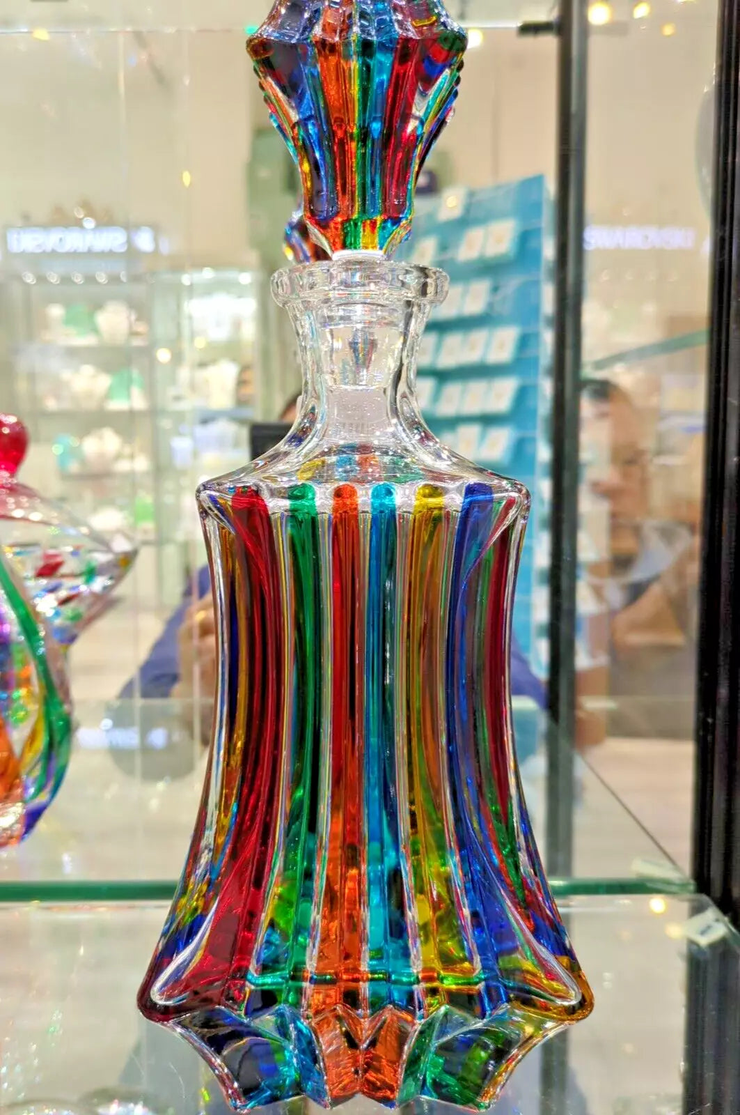 A tall, multi-colored Murano glass decanter with a narrow neck, displayed on a glass surface.