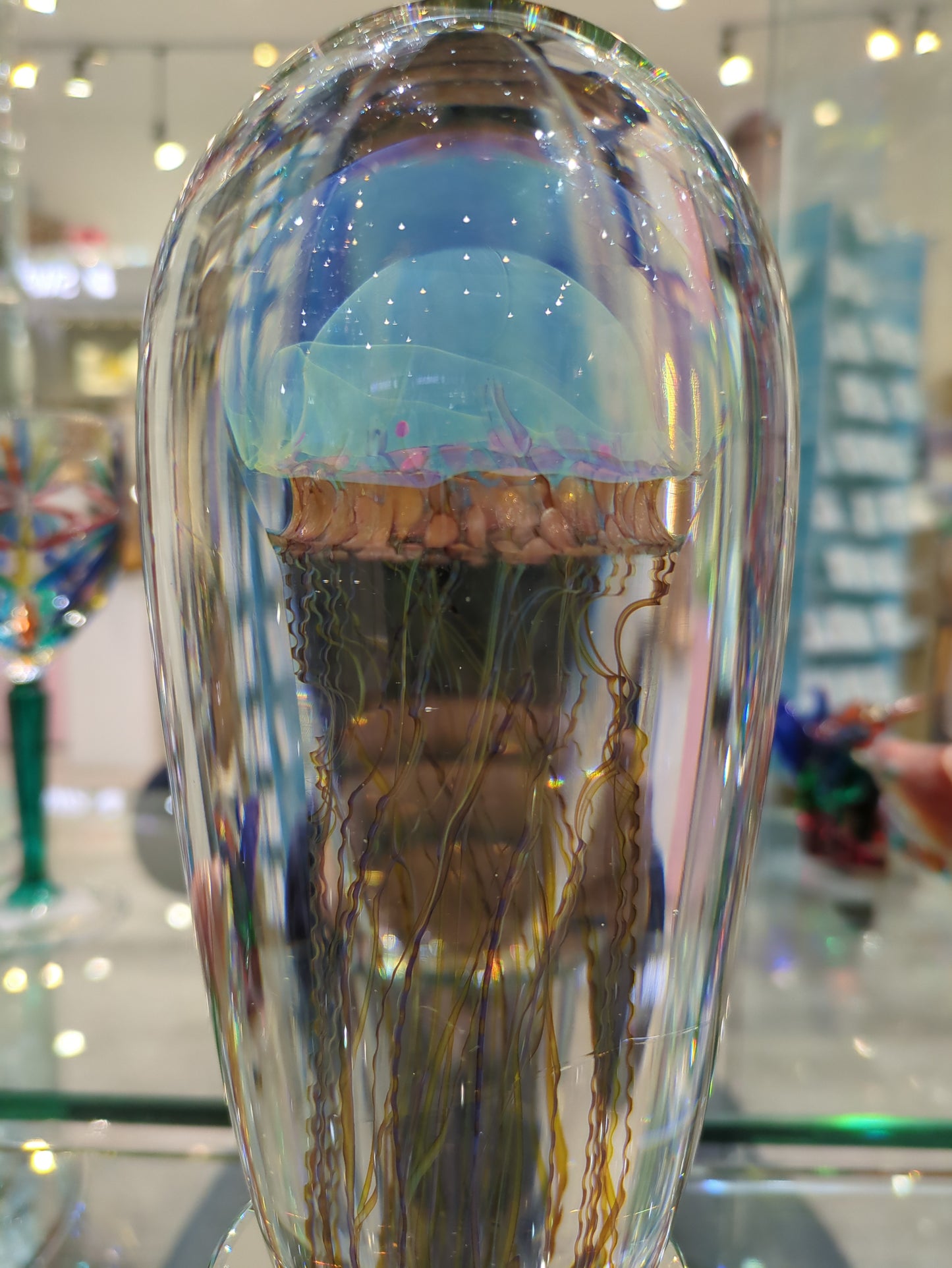 A Satava art glass jellyfish sculpture with a brown and gold band and colorful floating tentacles, displayed on a light base.
