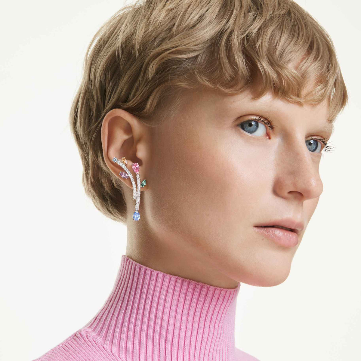 Model with short blonde hair wearing the Ariana Grande x Swarovski multicolored crystal drop earrings and a pink ribbed turtleneck.