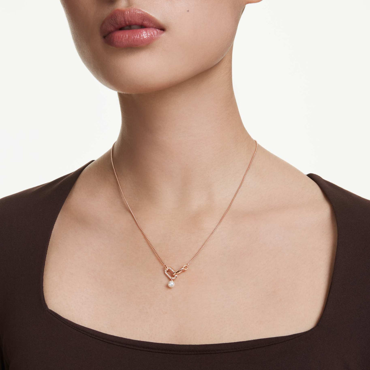Model wearing the Swarovski Constella pendant necklace with a rose gold-tone chain and pearl drop, shown over a dark brown top.