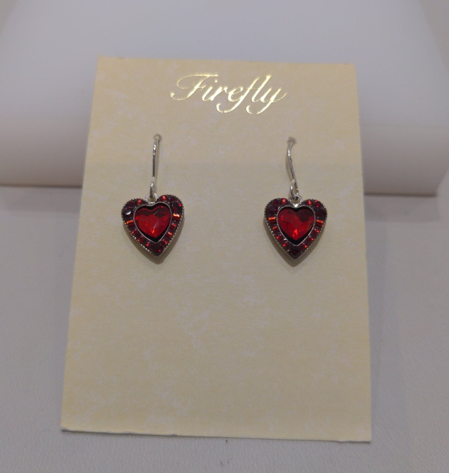 A pair of silver wire earrings with red heart-shaped embellishments, presented against a beige background in a clear case.
