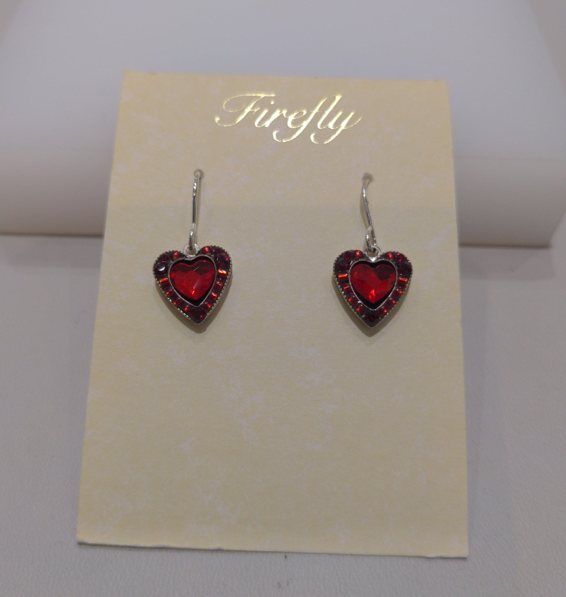A pair of silver wire earrings with red heart-shaped embellishments, presented against a beige background in a clear case.