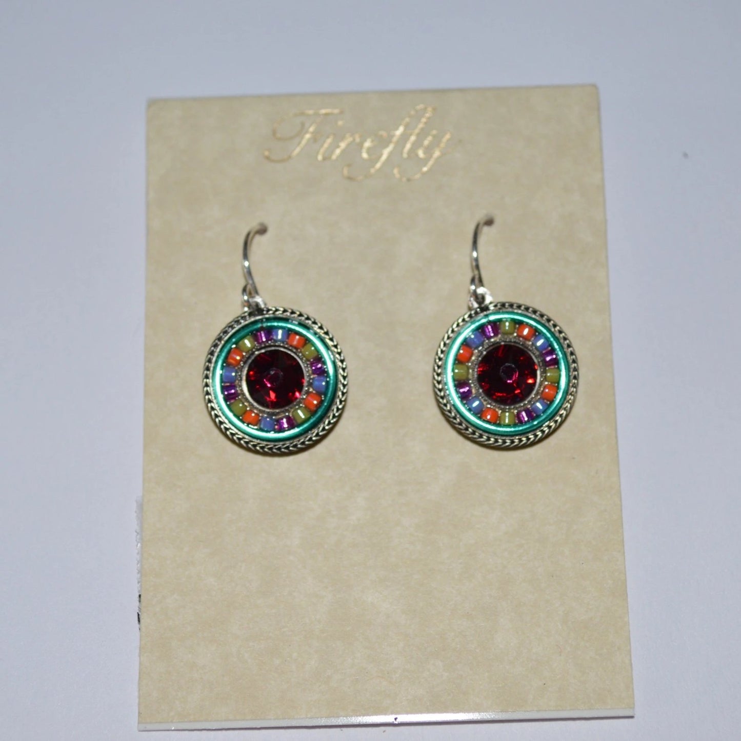 A pair of Firefly Jewelry earrings displayed on a card, featuring multicolored round pendants with intricate embellishments and a central red crystal.