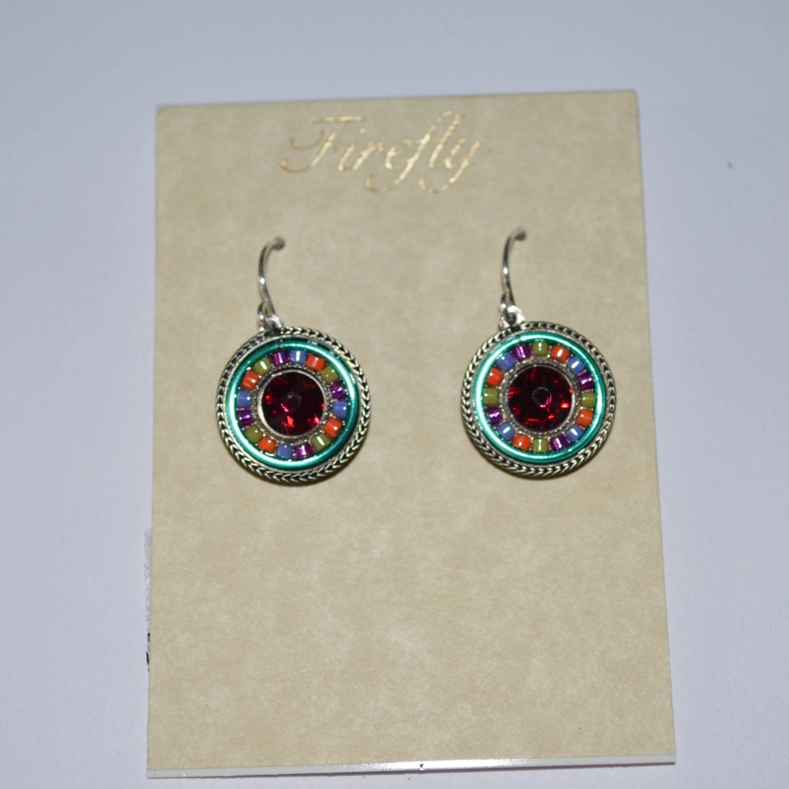 A pair of Firefly Jewelry earrings displayed on a card, featuring multicolored round pendants with intricate embellishments and a central red crystal.