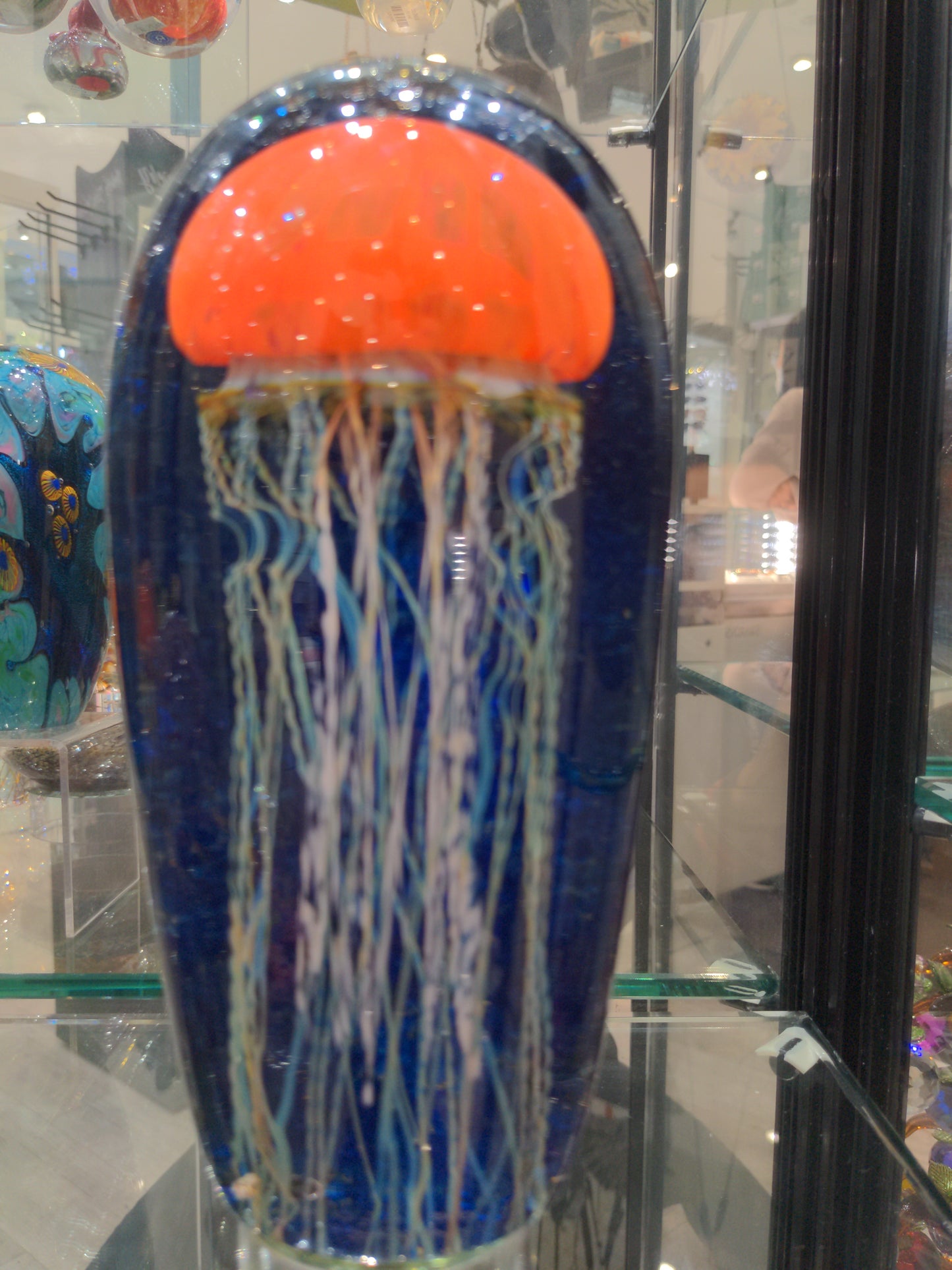 Satava art glass piece featuring an orange jellyfish with a blue background and white tentacles, displayed in a case.