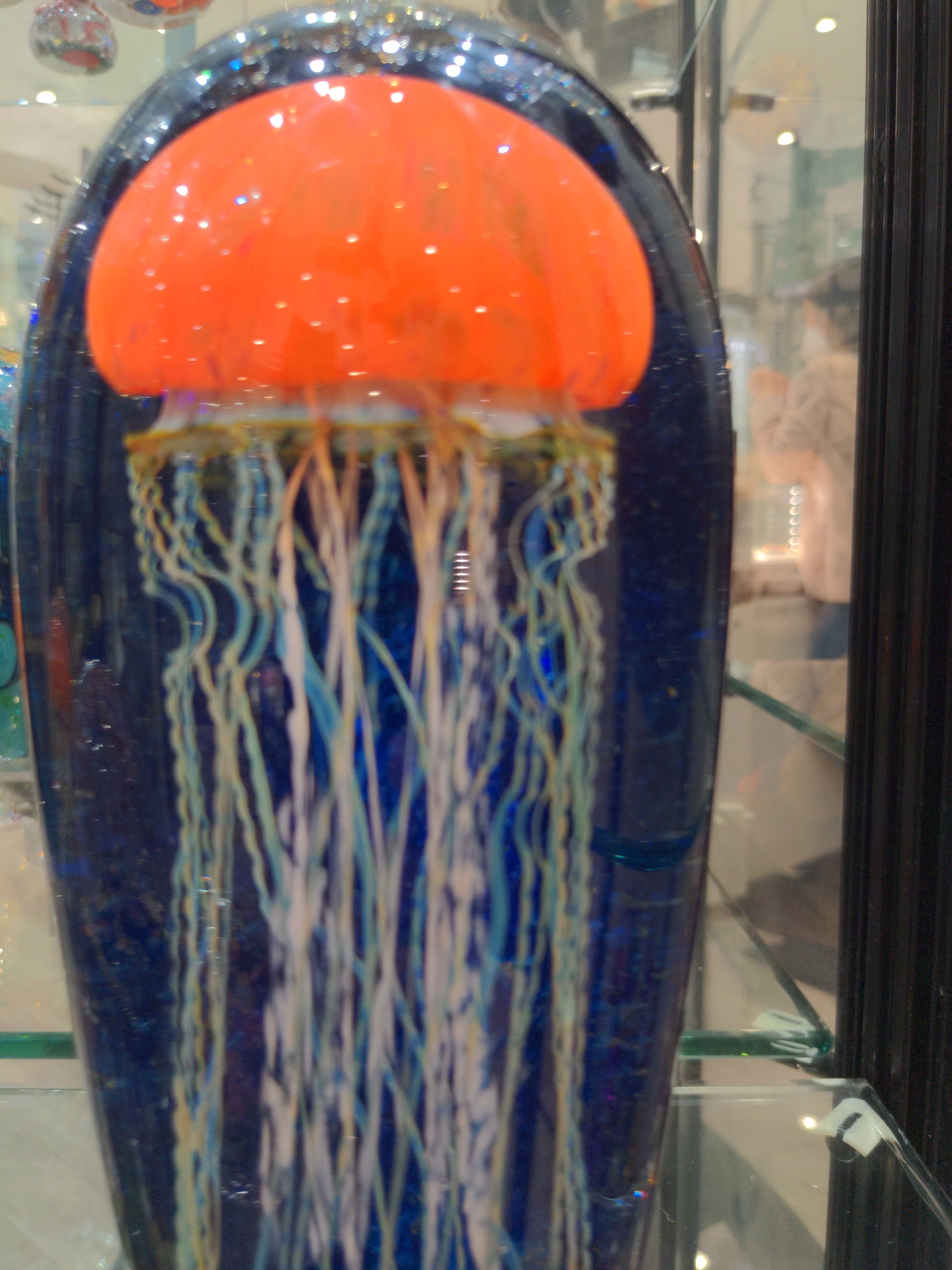 Satava art glass piece featuring an orange jellyfish with a blue background and white tentacles, displayed in a case.