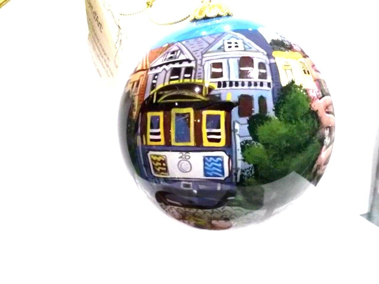 A round decorative ornament depicting a stylized San Francisco scene with the cable car on the side, placed in a green-trimmed box.