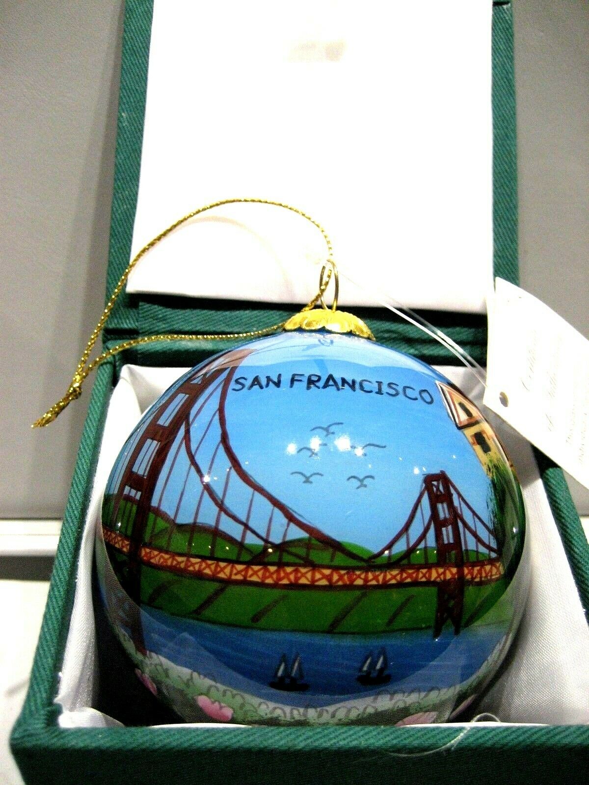 A round decorative ornament depicting a stylized San Francisco scene with the Golden Gate Bridge, placed in a green-trimmed box.